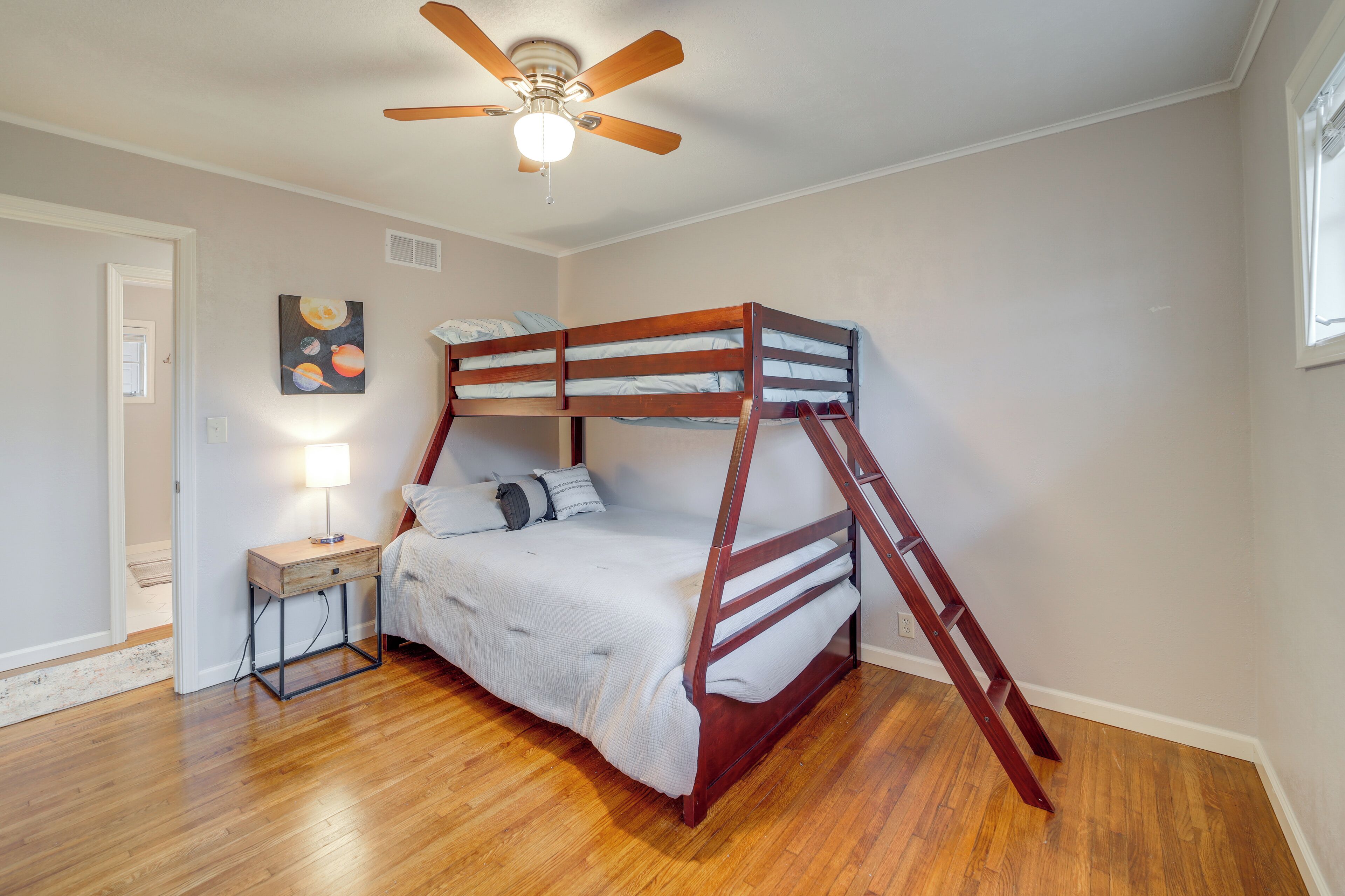 A bunk bed with a ladder, a bedside table with a lamp, and a ceiling fan in a room with wooden flooring and a door.