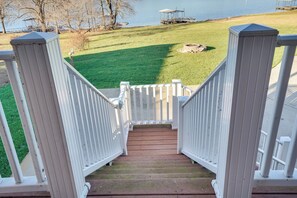 Un escalier en bois avec des rampes blanches mène à une zone herbeuse où se trouvent un quai et, au loin, une étendue d’eau.
