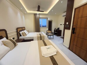 A hotel room with two beds, a desk, a chair, a ceiling fan, and a window with curtains. - Hare Krishna Stays (Varanasi)