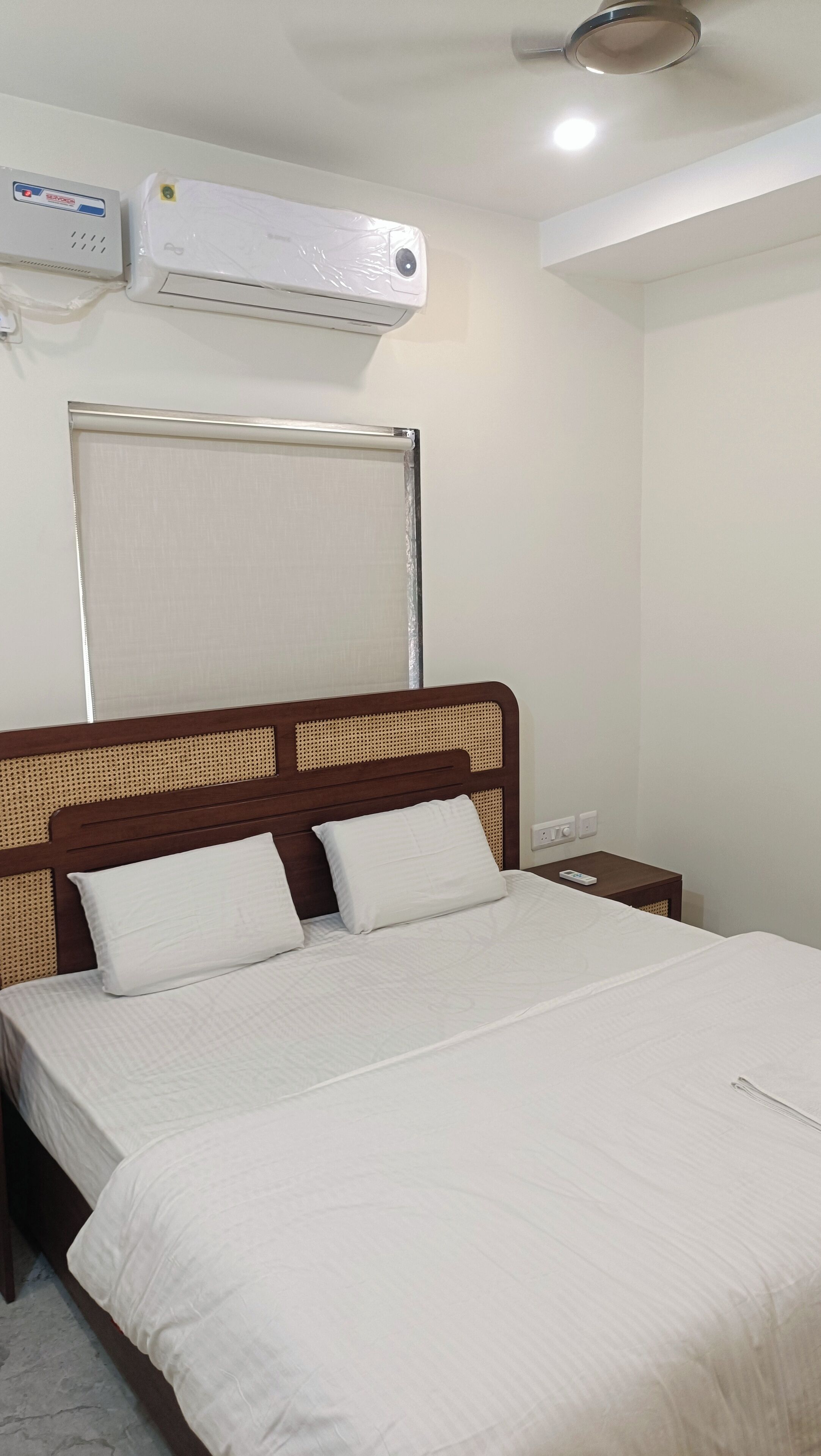Una cama bien hecha con sábanas blancas, cabecero de madera con respaldo tejido, aire acondicionado montado en la pared y una mesita de noche con lámpara.