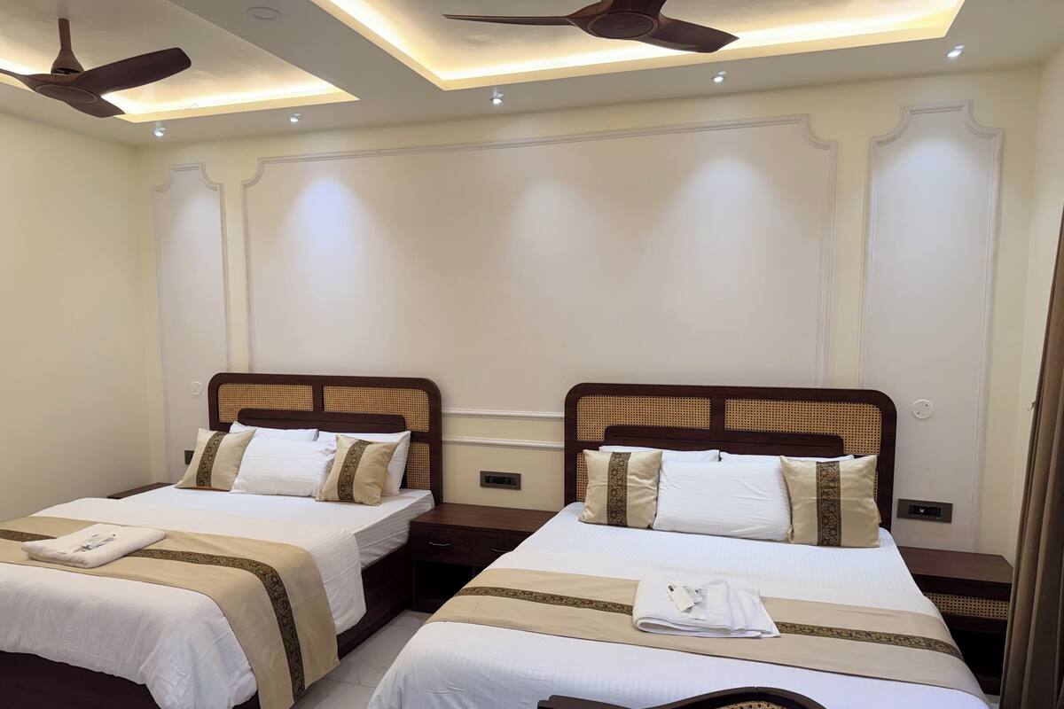A hotel room with two beds, each with a wooden headboard, white bedding, and a beige accent strip. There are ceiling fans, recessed lighting, and a small table with a chair.