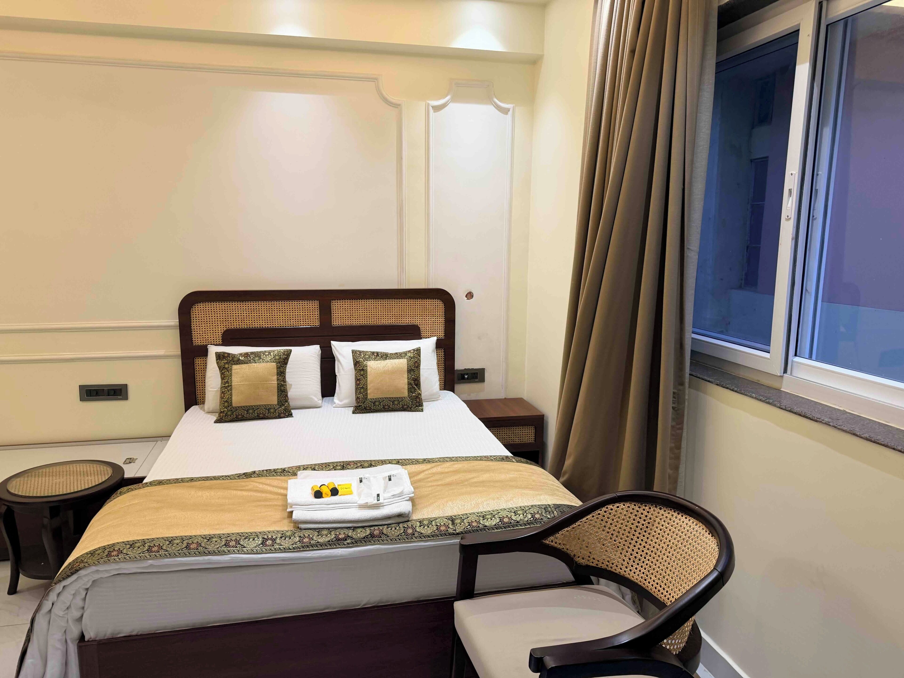 A neatly made bed with two pillows, a bedside table, a chair, and a window with curtains.