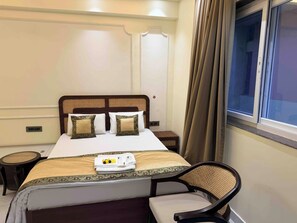 A neatly made bed with two pillows, a bedside table, a chair, and a window with curtains. - Hare Krishna Stays (Varanasi)