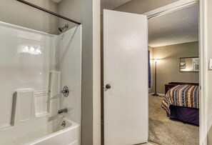 Un baño con bañera, ducha y una puerta que da a un dormitorio con cama y mesita de noche.