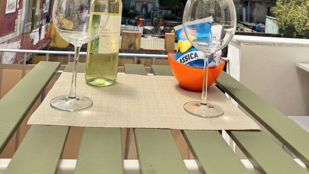 A balcony with a table set for two, featuring a bottle of white wine and two glasses. A basket with snacks and a bag of chips is also visible. The background includes a view of a historic stone building and a tree. - House named Borghesia in the heart of the Sanità district of Naples (Napoli)