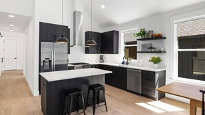Une cuisine moderne avec des meubles de couleur sombre, des appareils électroménagers en acier inoxydable, un îlot central doté de tabourets et un coin repas avec une table en bois.
