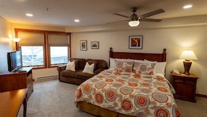 A bedroom with a bed, a desk, a chair, a ceiling fan, and a window with blinds. - 1307b - King Standard Eagle Springs West Hotel Room (Solitude)