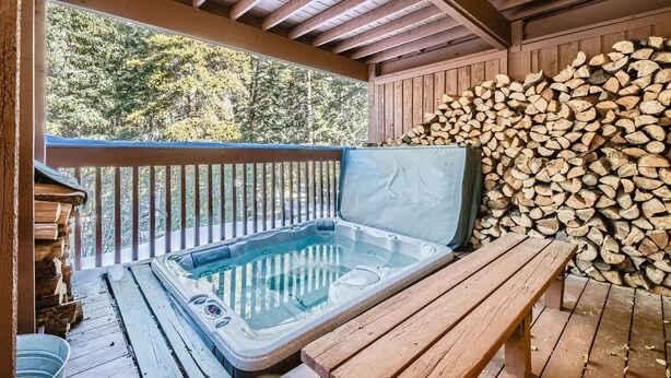 Un jacuzzi en una terraza de madera con vista a árboles, una pila de leña y un balde de metal.
