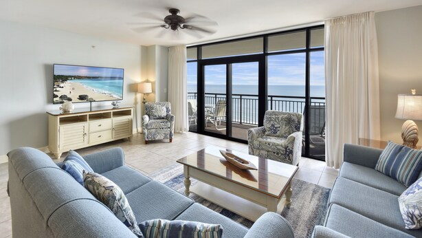 A spacious living room with a large window overlooking a balcony, a ceiling fan, and a flat-screen TV on the wall.