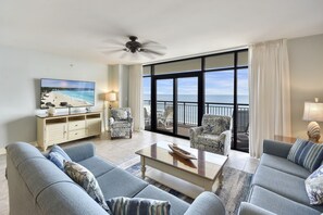 Een ruime woonkamer met een groot raam dat uitkijkt op een balkon, een plafondventilator en een flatscreen televisie aan de muur.