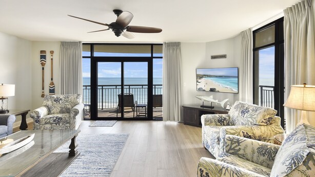 A spacious living room with a large window offering a view of the ocean, a ceiling fan, and a flat-screen TV mounted on the wall. - Southwind 806 5 Bedroom Condo (Myrtle Beach)