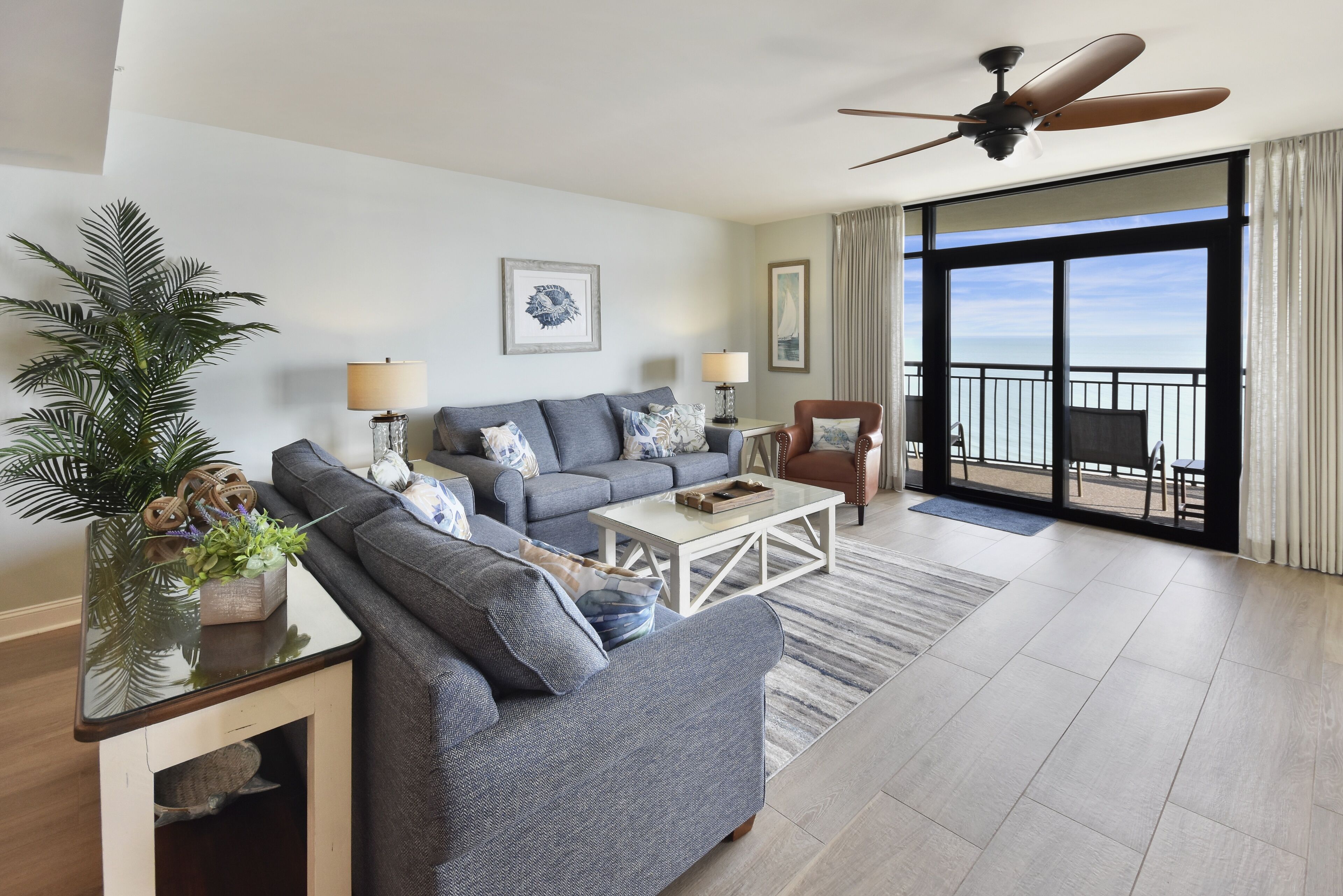 A modern living room with a grey sofa, a coffee table, and a ceiling fan. There is a large window with a view of the outdoors.