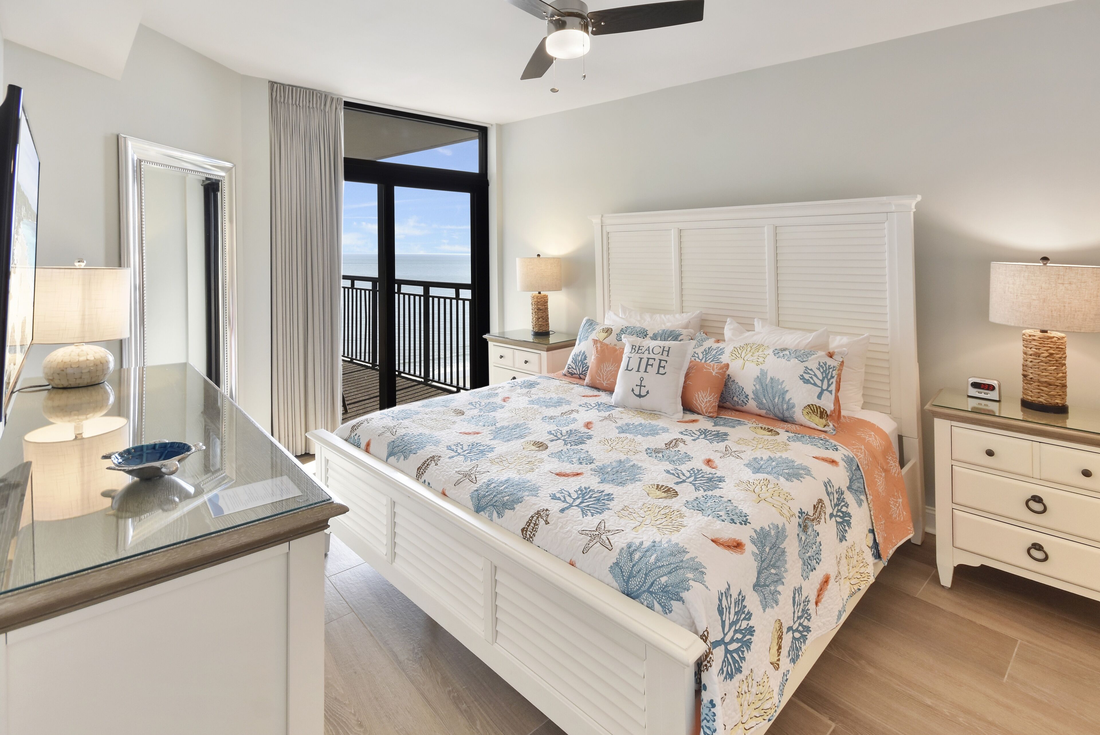 A bedroom with a large bed, a nightstand, a dresser, a mirror, and a balcony with a view.