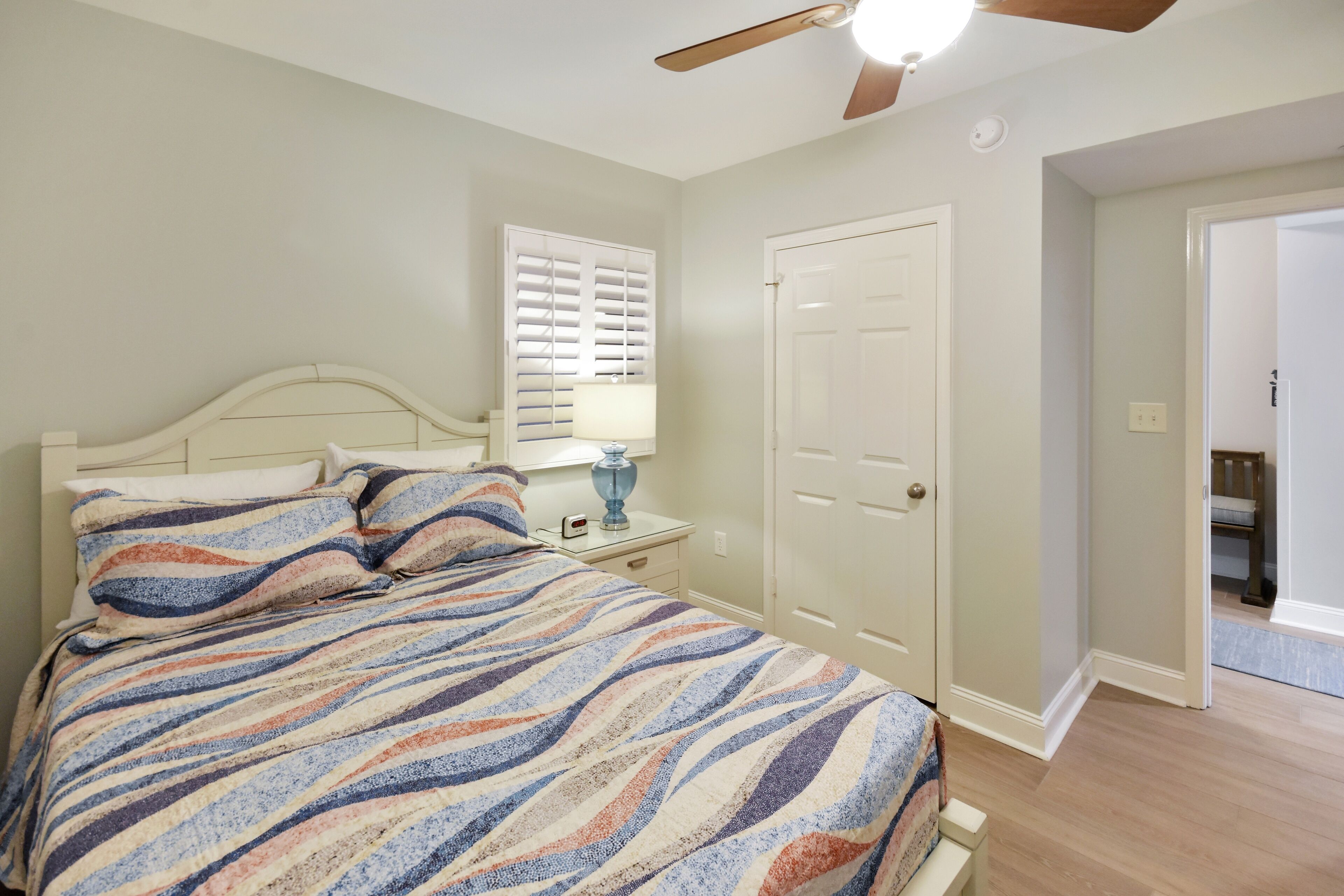 Une chambre à coucher avec un lit, une table de chevet, une lampe, un ventilateur de plafond et une porte.