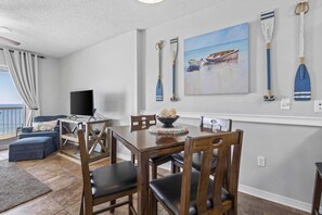 A dining area with a wooden table and chairs, a television, and a painting of boats on the wall. - Caribbean 502 1 Bedroom Condo (Gulf Shores)