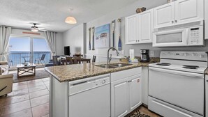 Une cuisine avec des armoires blanches, un comptoir en granit, un four à micro-ondes et une cuisinière. On y trouve également un coin repas avec une table et des chaises, et une vue sur l’océan par une porte-fenêtre coulissante.