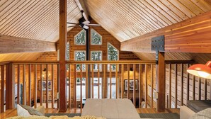 A cozy cabin interior with a sloped ceiling, wooden beams, a bed, a sofa, and large windows offering a view of trees.