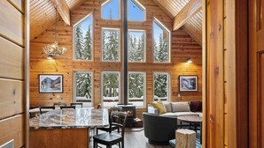 A log cabin interior with a large dining area, a fireplace, and multiple windows offering views of snow-covered trees.
