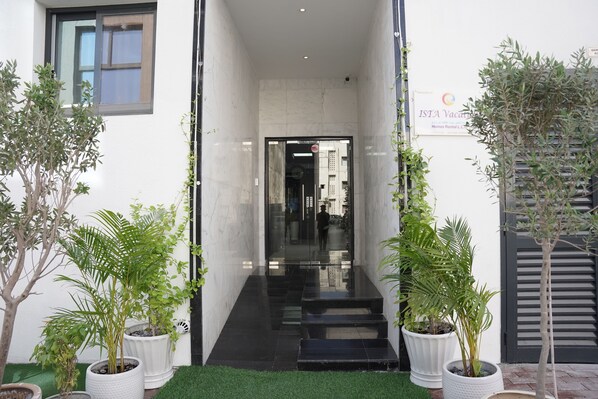 Welcome to the entrance of our hotel, featuring a sleek glass door and steps leading up to a threshold, surrounded by potted plants.