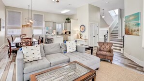 A spacious living room with a gray sectional sofa, a glass coffee table, and a dining area with a wooden table and chairs. A staircase leads to an upper level. - Luxurious gulf side living with Golf Cart! + Free Dolphin Cruise! (Santa Rosa Beach)