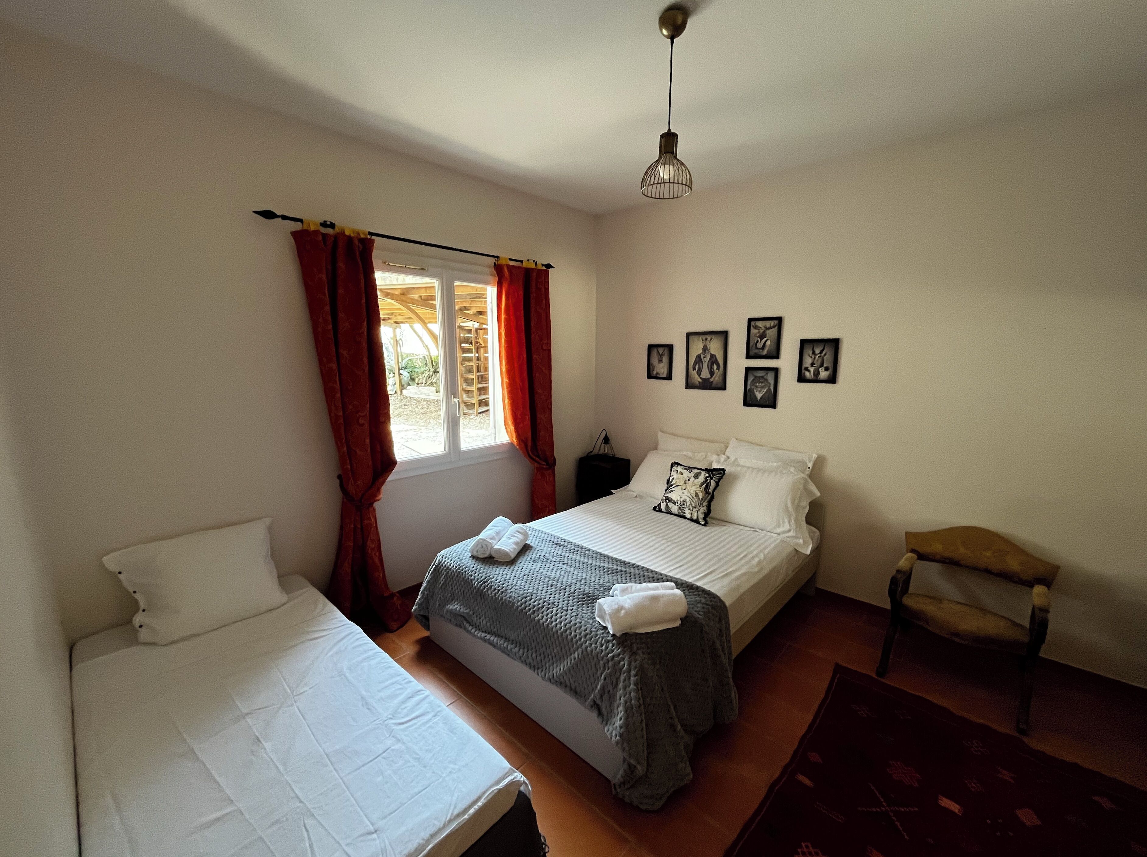 A bedroom with two beds, a window with red curtains, a bedside table, and wall art.