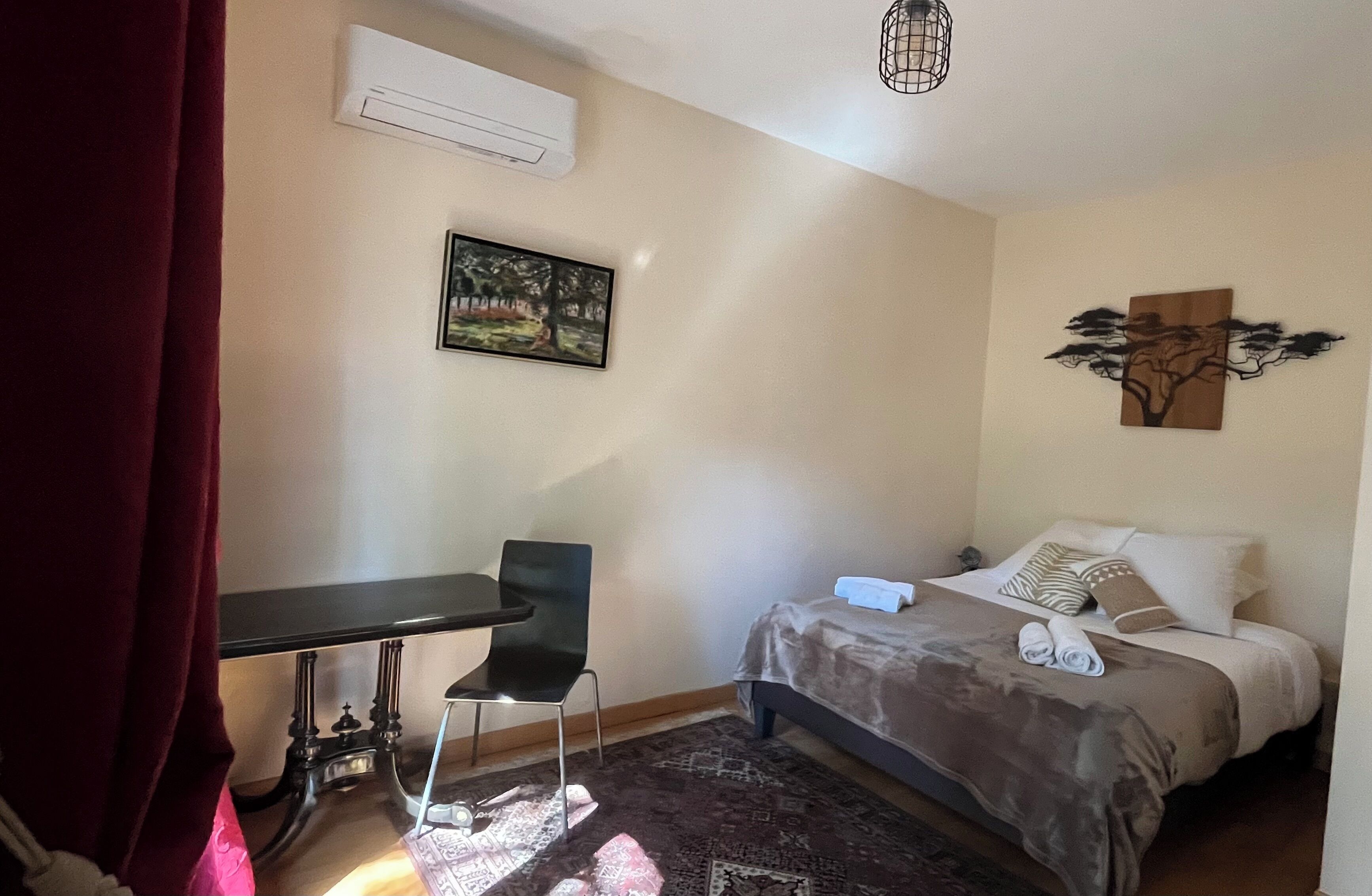 A bedroom with a bed, a desk, a chair, an air conditioner, and a painting on the wall.