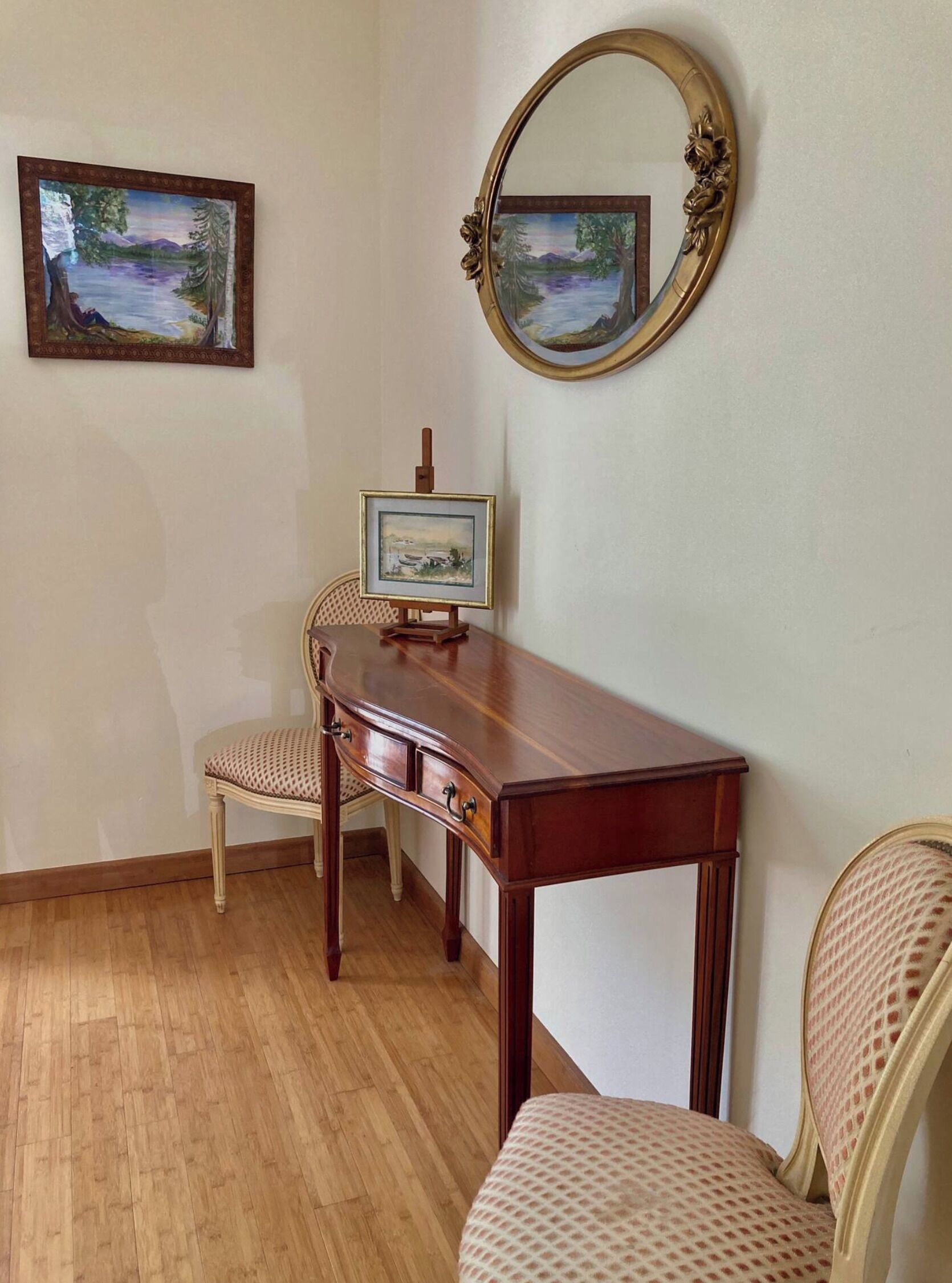 Une pièce avec un bureau en bois, deux chaises, un petit chevalet sur lequel est posé un tableau, et un miroir accroché au mur.