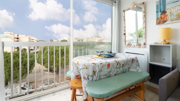 A balcony with a view of buildings and trees, a table with a colorful tablecloth, a bench, and a lamp on a shelf.