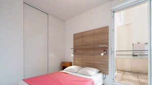 A modern bedroom with a bed, a nightstand, a wooden headboard, and a sliding glass door leading to an outdoor area. - Bright, air-conditioned appartement 4 ppl,  close to the sea (Le Grau-du-Roi)