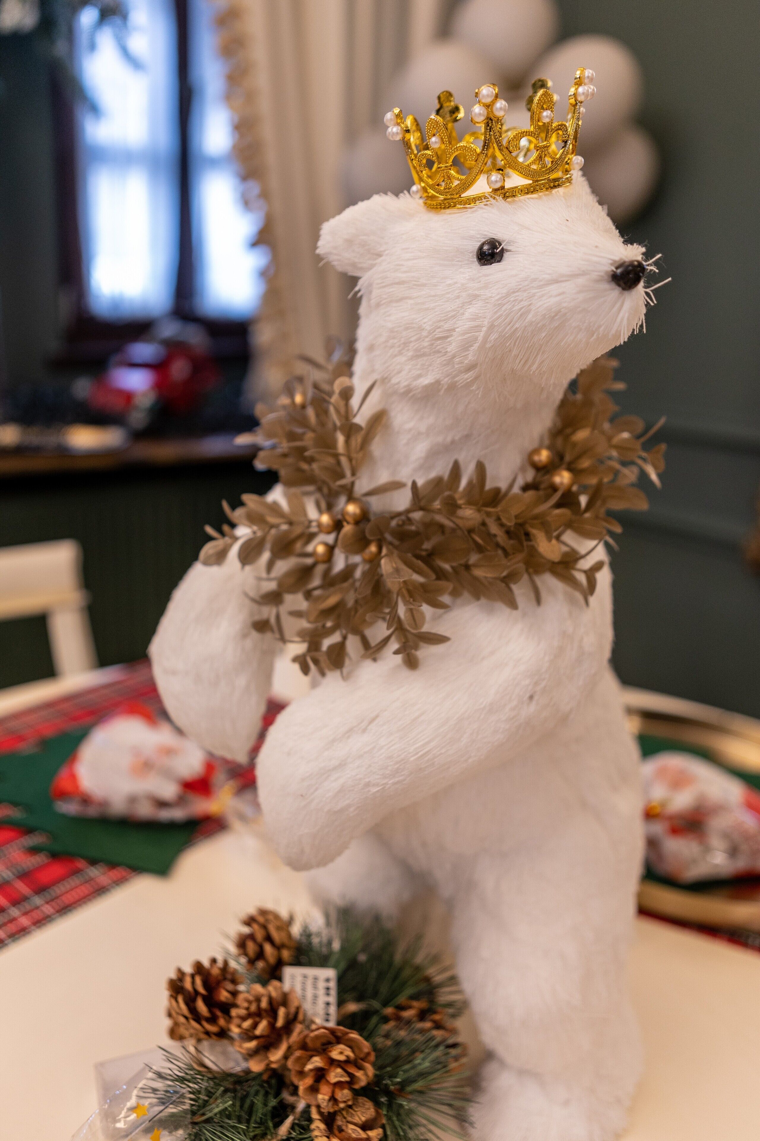A white bear figurine with a golden crown and a garland, placed on a table with a plaid tablecloth and pine cones.