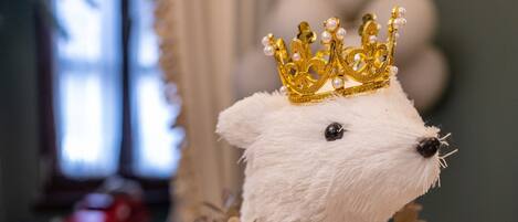 A white bear figurine with a golden crown and a garland, placed on a table with a plaid tablecloth and pine cones.