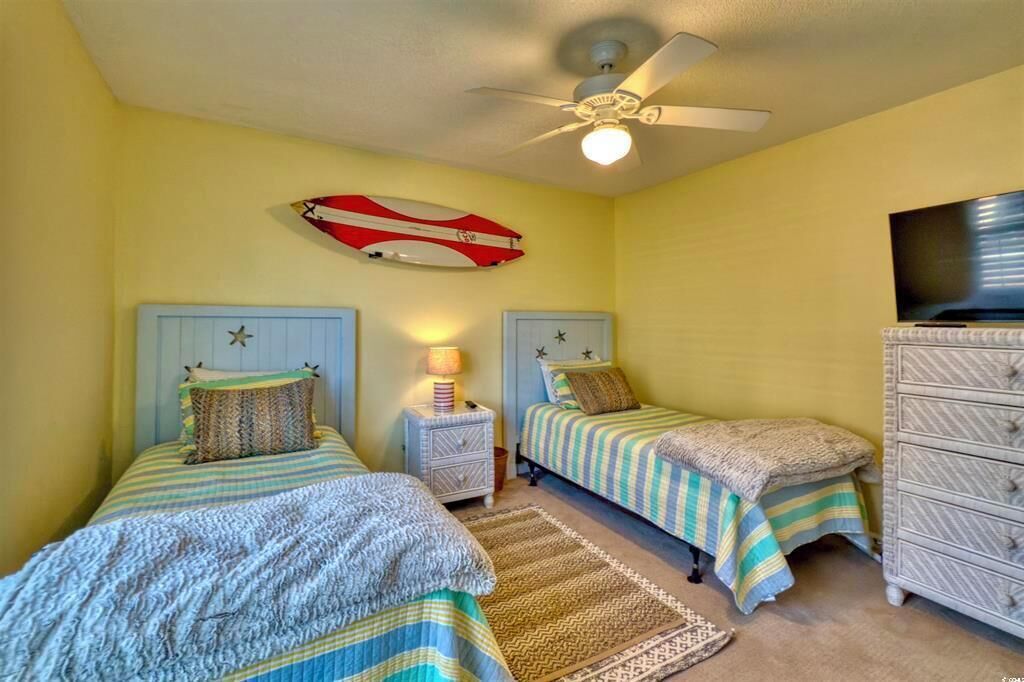 Une chambre avec deux lits, un ventilateur de plafond, une télévision et une planche de surf décorative accrochée au mur.