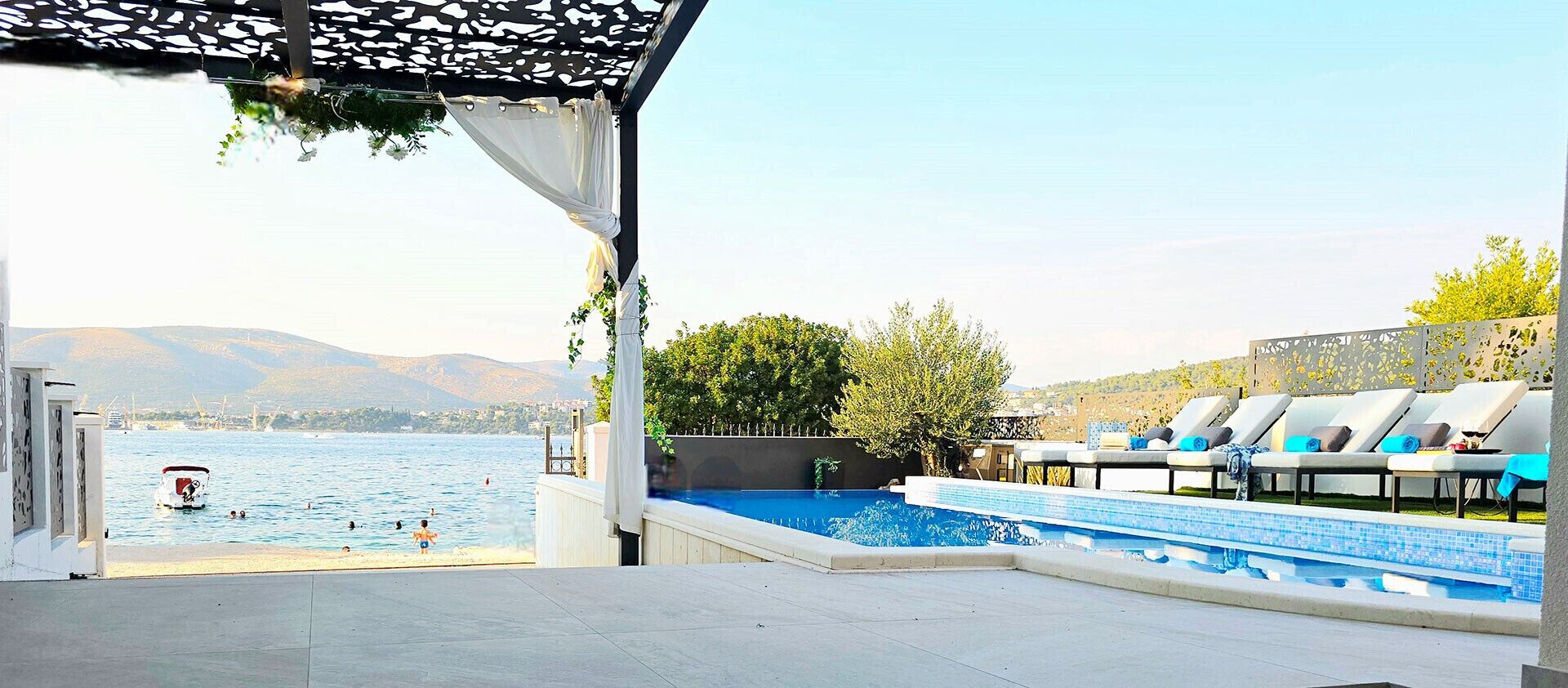 A pool area with lounge chairs, a view of the sea, and mountains in the background.