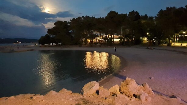 Ein Strand bei Dämmerung mit ruhigem Wasser, das Lichter, Bäume und eine ferne Küste spiegelt.