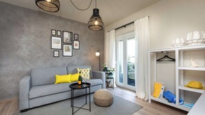 Una sala de estar moderna con un sofá gris, un banquito redondo, una mesita negra con una botella y vasos, y un estante blanco con sombreros y bolsos.