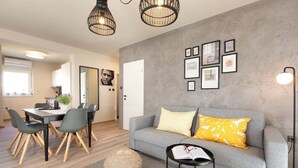 Una sala de estar moderna con un sofá gris, un otoman redondo, un área de comedor con una mesa y sillas, y un pasillo que conduce a una cocina.