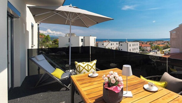 Un balcón con una mesa de madera, una sombrilla blanca y vista al mar y edificios.