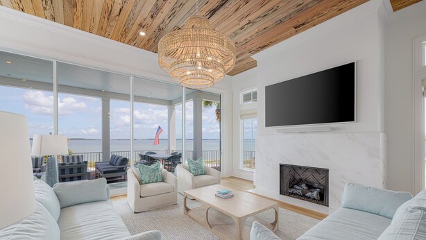 A spacious living room with a large window overlooking the sea, a fireplace, and a modern chandelier. - Luxury Sound-View Escape with Private Pool, Hot Tub, and Stunning Amenities! (Pensacola Beach)