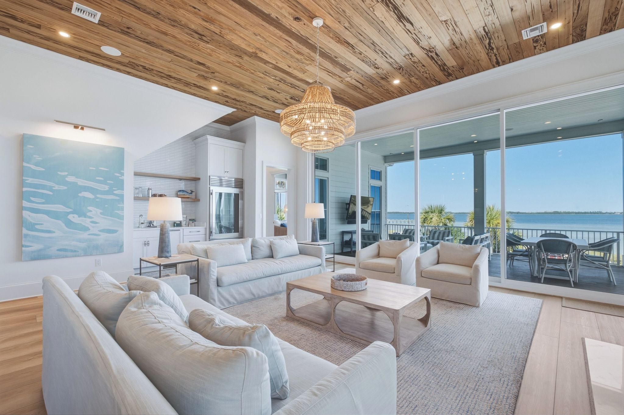 A spacious living room with a large window overlooking a body of water, a chandelier, and a modern kitchen in the background.