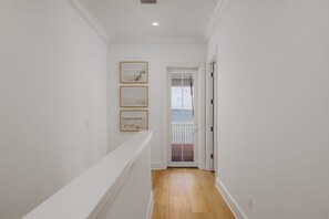A hallway with white walls, wooden flooring, and a door leading to a balcony with a view of the sea.