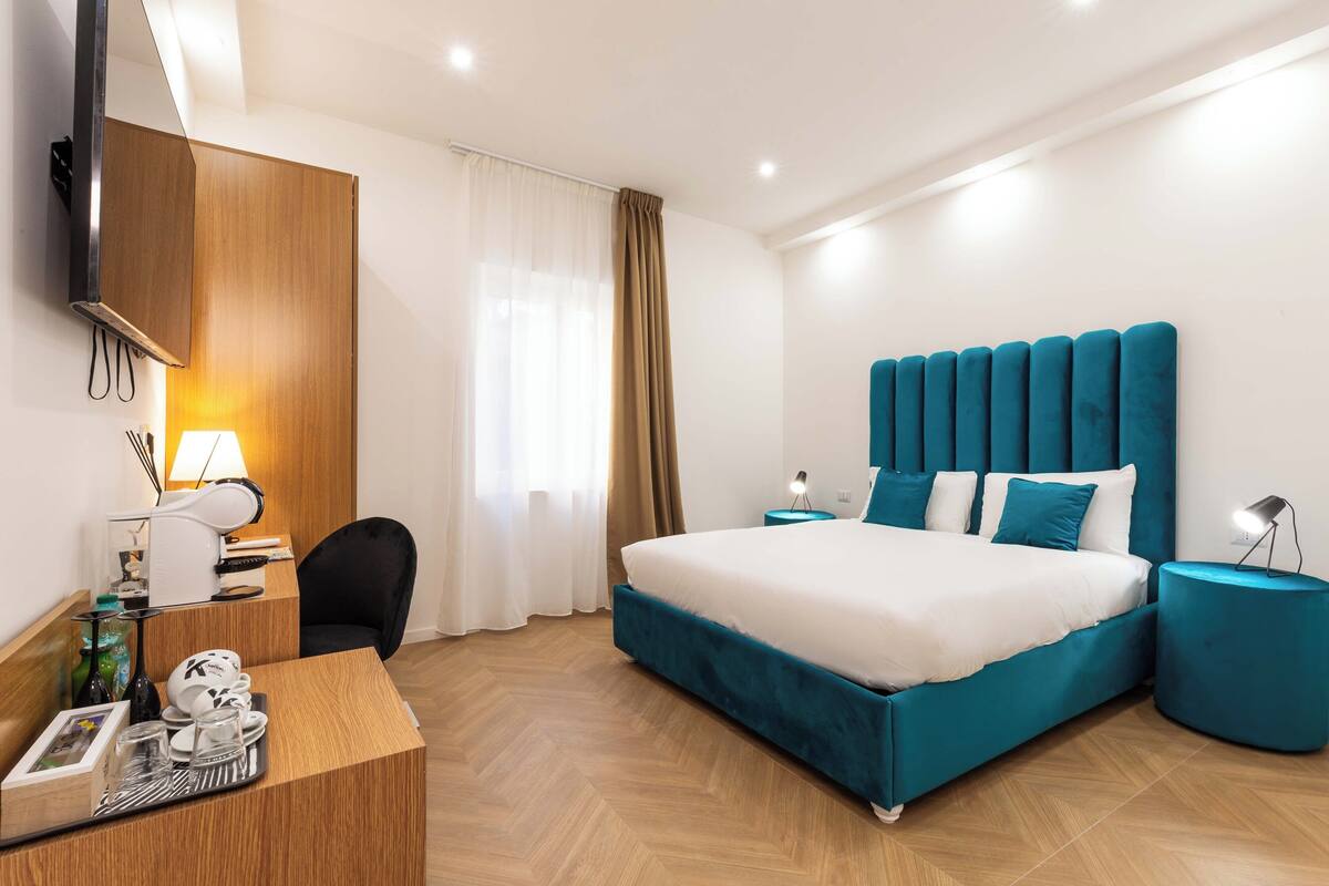 A modern hotel room with a large bed, a wooden desk, a chair, a TV mounted on the wall, and a window with curtains.