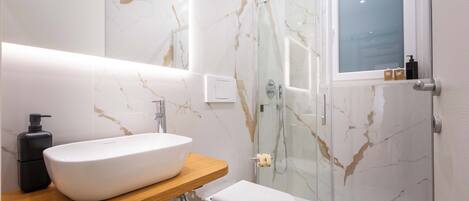 A modern bathroom with a marble backsplash, a white sink, a glass shower enclosure, and a wooden vanity.