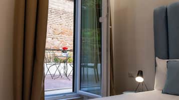 Un dormitorio con cama, una mesita de noche con lámpara, ventana con cortinas y vistas a un patio exterior con mesa y sillas.