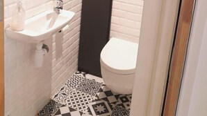 Une petite salle de bain moderne avec un tableau noir sur lequel est écrit « Bienvenue » et trois cœurs. Elle comprend un lavabo blanc, des toilettes et des carreaux de sol à motifs.