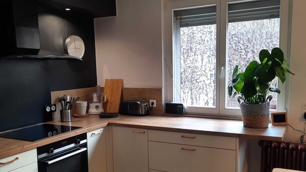 Une cuisine moderne avec un revêtement mural noir, des armoires blanches, un four encastré et une fenêtre donnant sur la forêt.