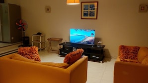 Una sala de estar con dos sofás naranjas, un televisor sobre un mueble bajo, un ventilador y un cuadro en la pared.