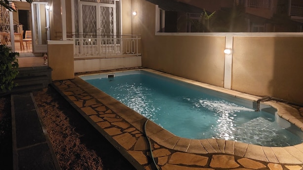 Una piscina con baldosas de piedra, una zona cubierta con barandilla y un edificio con balcones y ventanas.