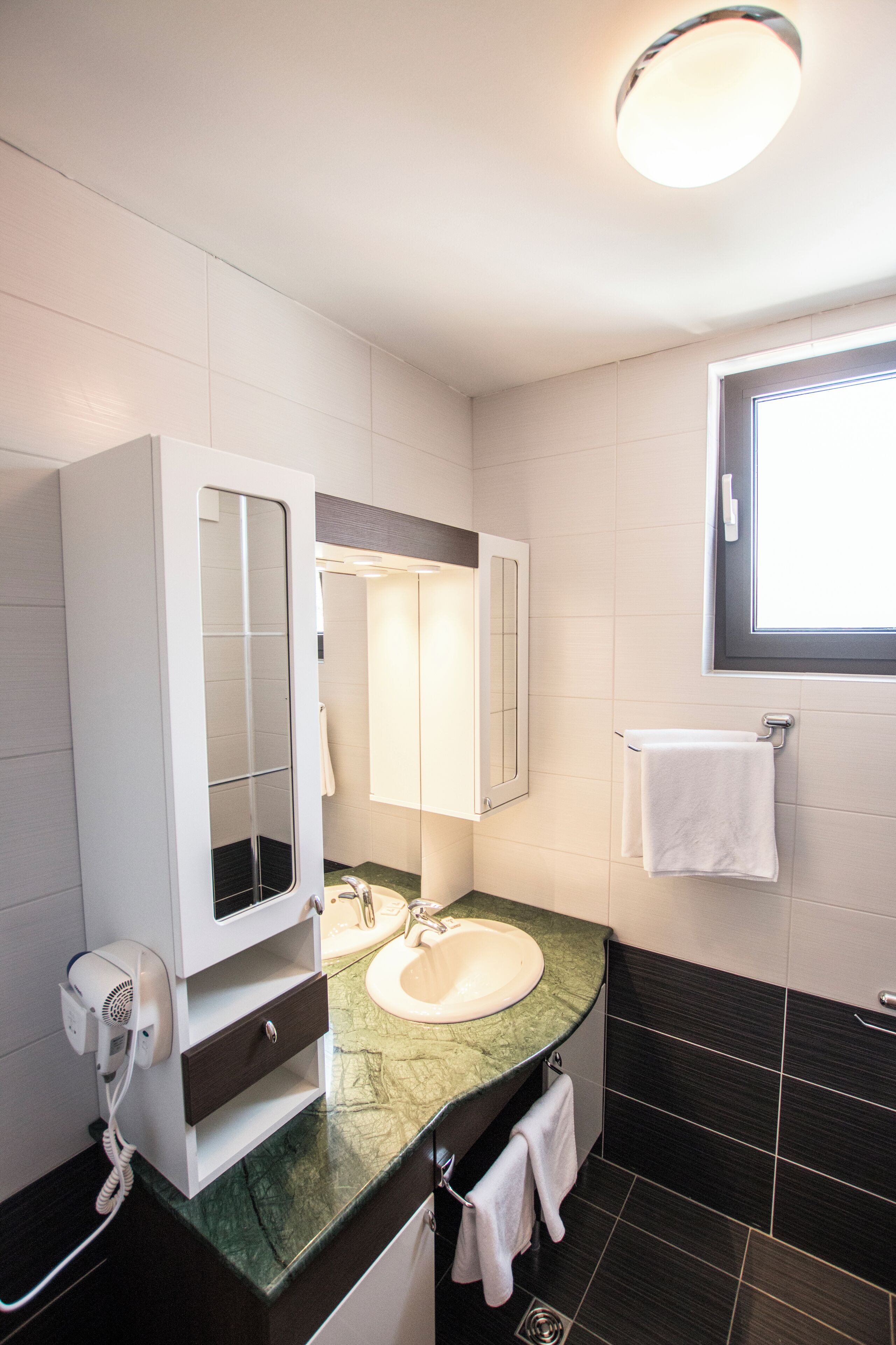 A modern bathroom with a white vanity, a large mirror, a sink, and a window above the mirror.