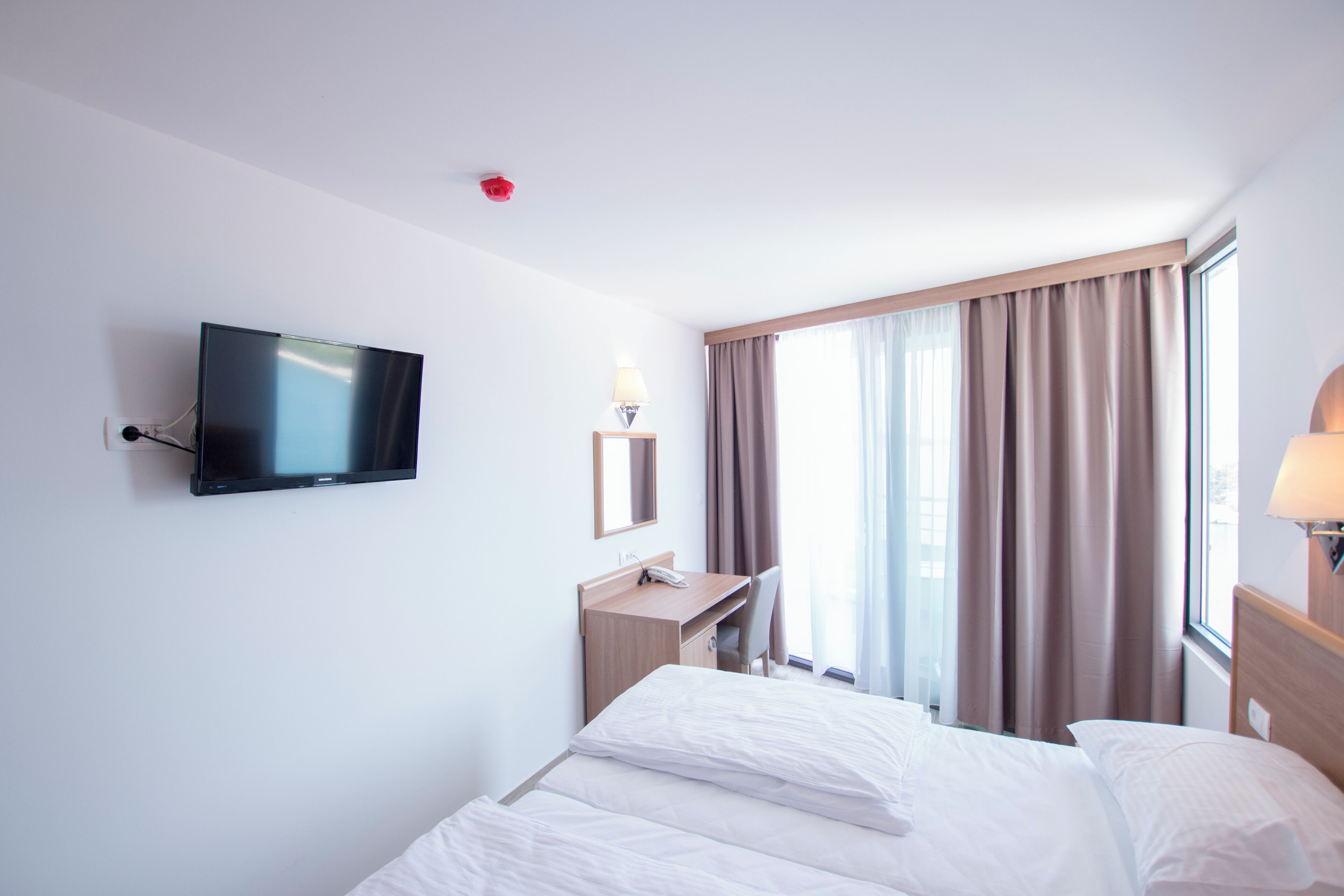 Quarto de hotel com cama, escrivaninha, cadeira, televisão e janela com cortinas.