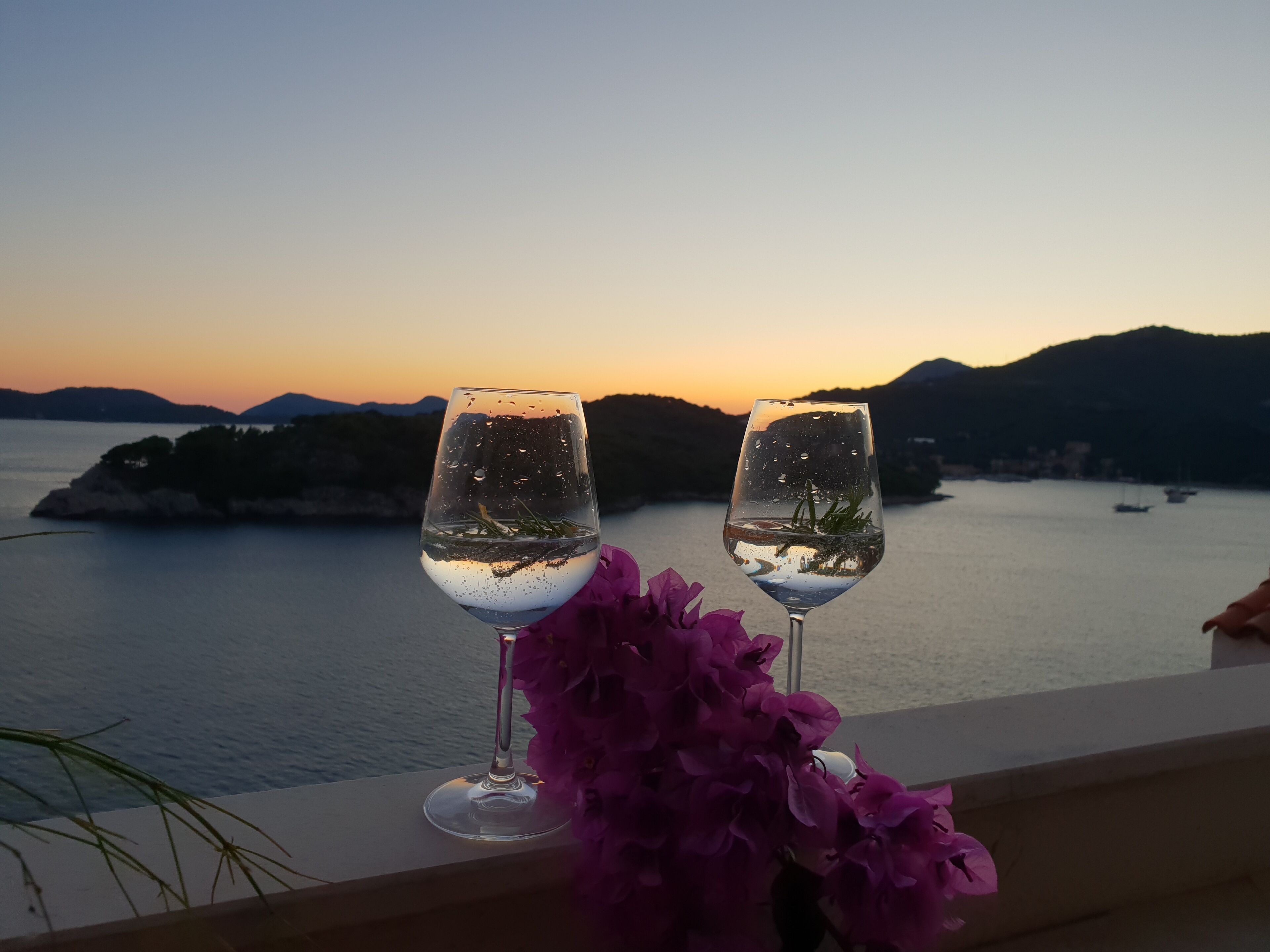 Two glasses of water with a flower on a balcony overlooking a body of water at sunset.